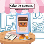 Cross stitch luggage tag kit for 'Calvin the Cappuccino' with a coffee-themed design on a table with a plate of pastries.