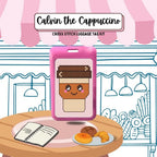 Cross stitch luggage tag kit for 'Calvin the Cappuccino' on a table with a notebook and pastries.