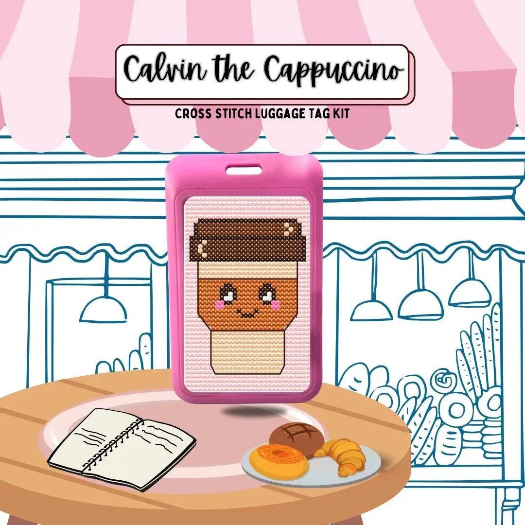 Cross stitch luggage tag kit for 'Calvin the Cappuccino' on a table with a notebook and pastries.