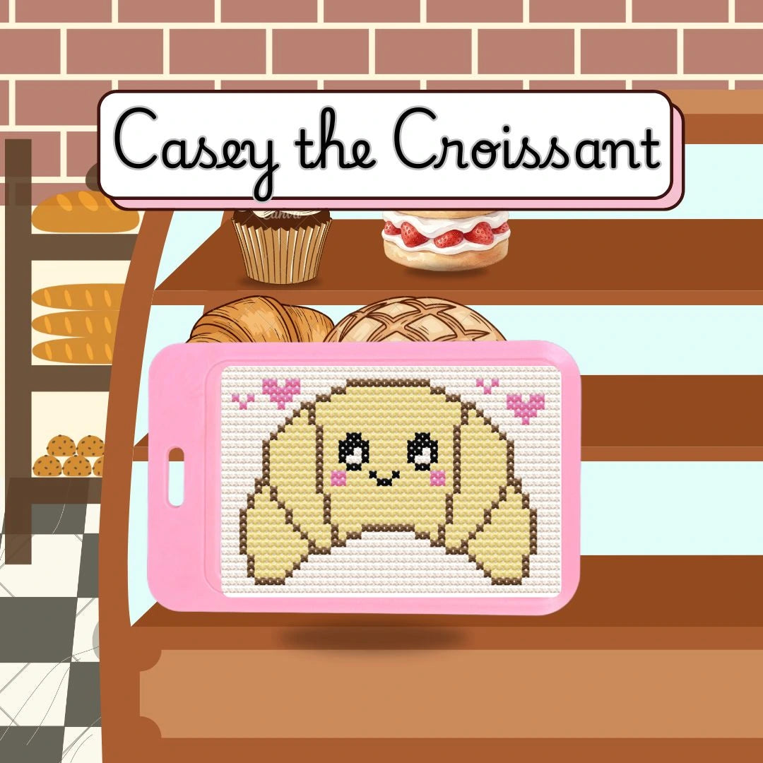 Cross-stitch pattern of a croissant with a smiling face on a pink hoop, set against a bakery background.