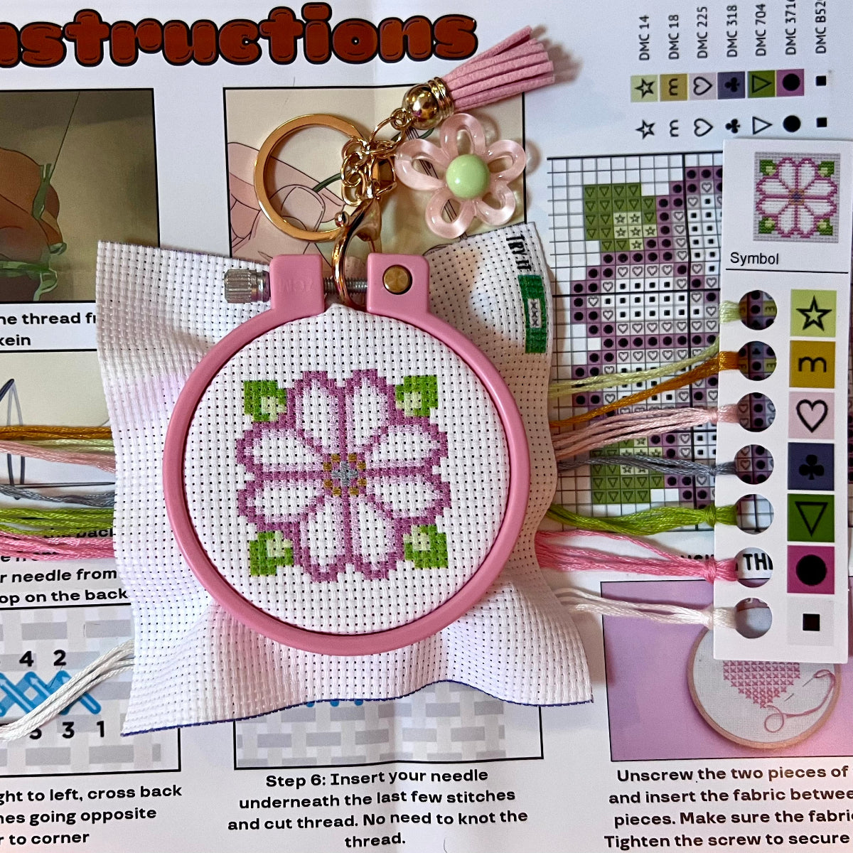 Cross-stitched keychain with pink hoop and floral design, surrounded by crafting materials and instructions.