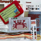 Cross-stitch kit with a crab pattern, threads, and instructions on a white background
